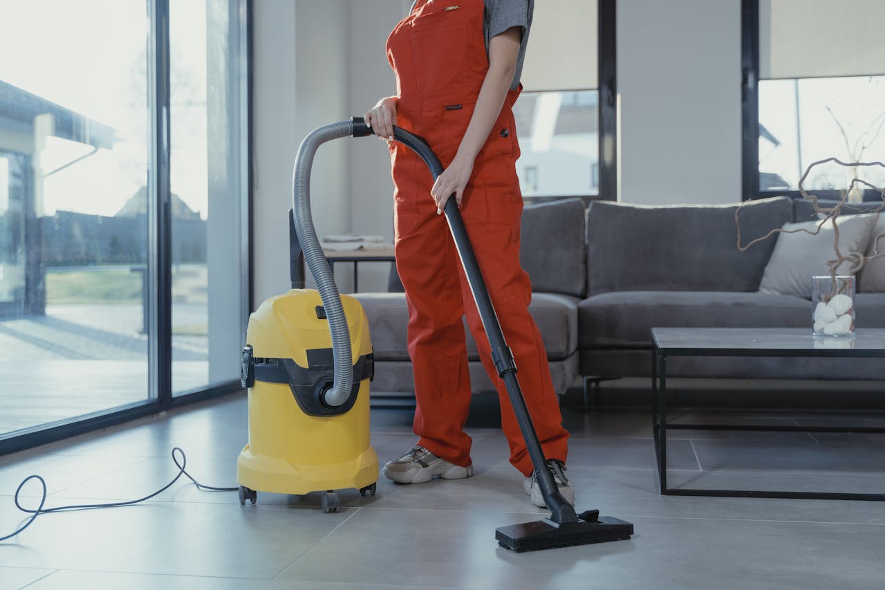 services-02 A professional cleaner in red coveralls vacuuming a modern living room with a yellow vacuum cleaner.